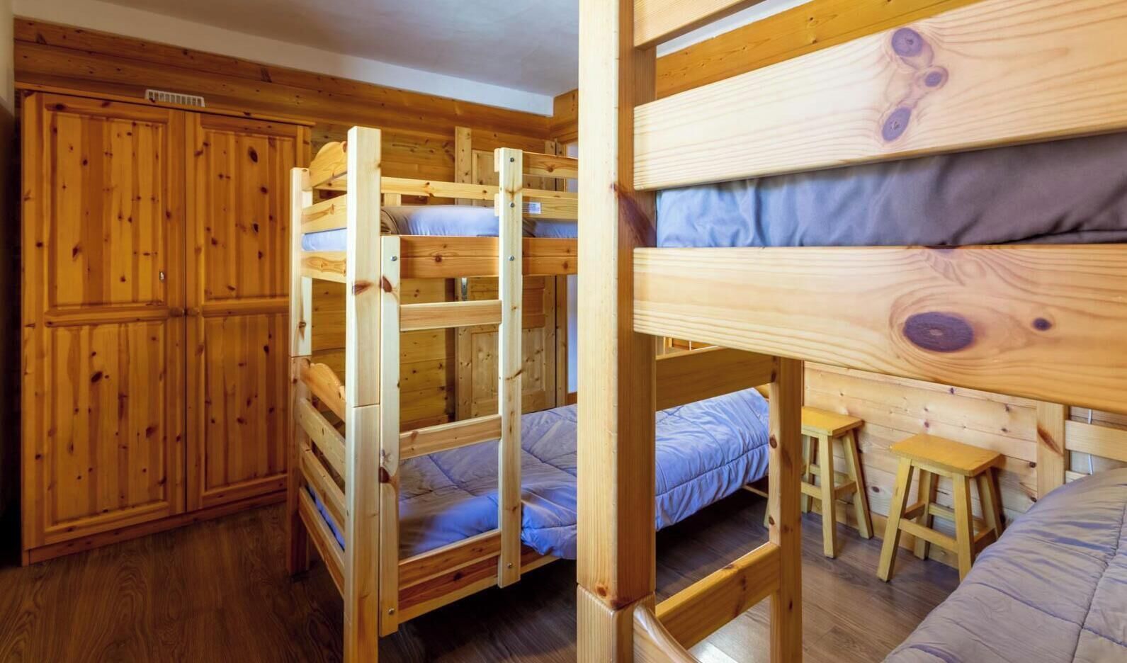Une chambre avec un lit superposé, une armoire en bois et un petit bureau avec des tabourets.