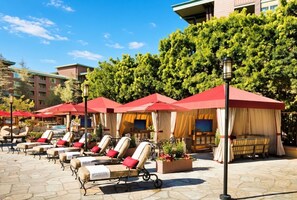 A poolside area with sun loungers and umbrellas, a stone floor, and a building in the background.