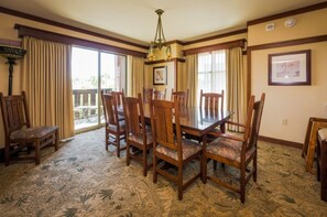 A dining area with a glass-top table surrounded by wooden chairs with cushioned seats, a patterned carpet, and a chandelier.