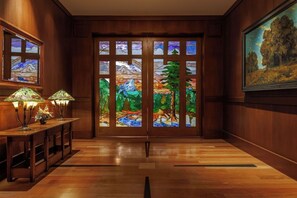 A room with wooden paneled walls, a wooden floor, a desk with two lamps, and a large stained glass window with a landscape view.