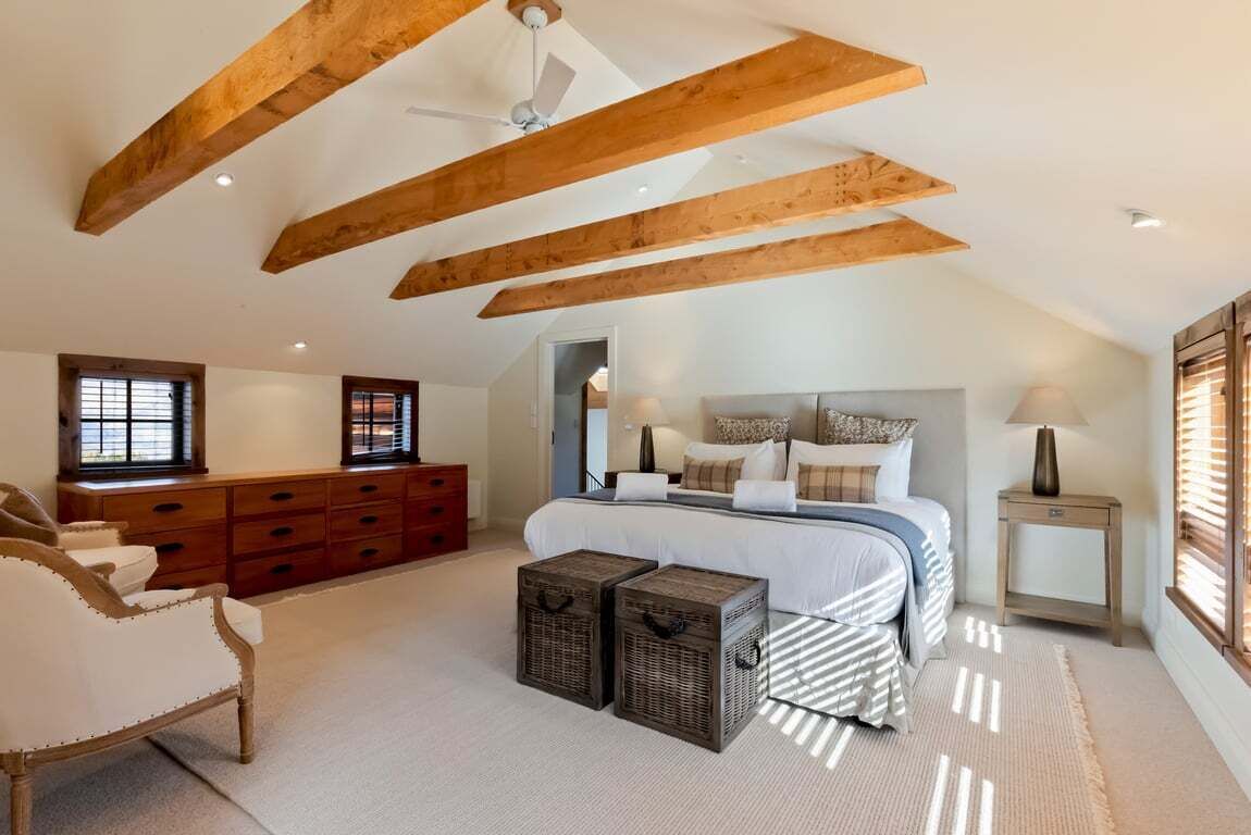 Ein Schlafzimmer mit schrägen Wänden, Holzbalken, einem Bett mit weißer Bettwäsche, einem Nachttisch, einer Lampe, einer Kommode und zwei Korbwannen.