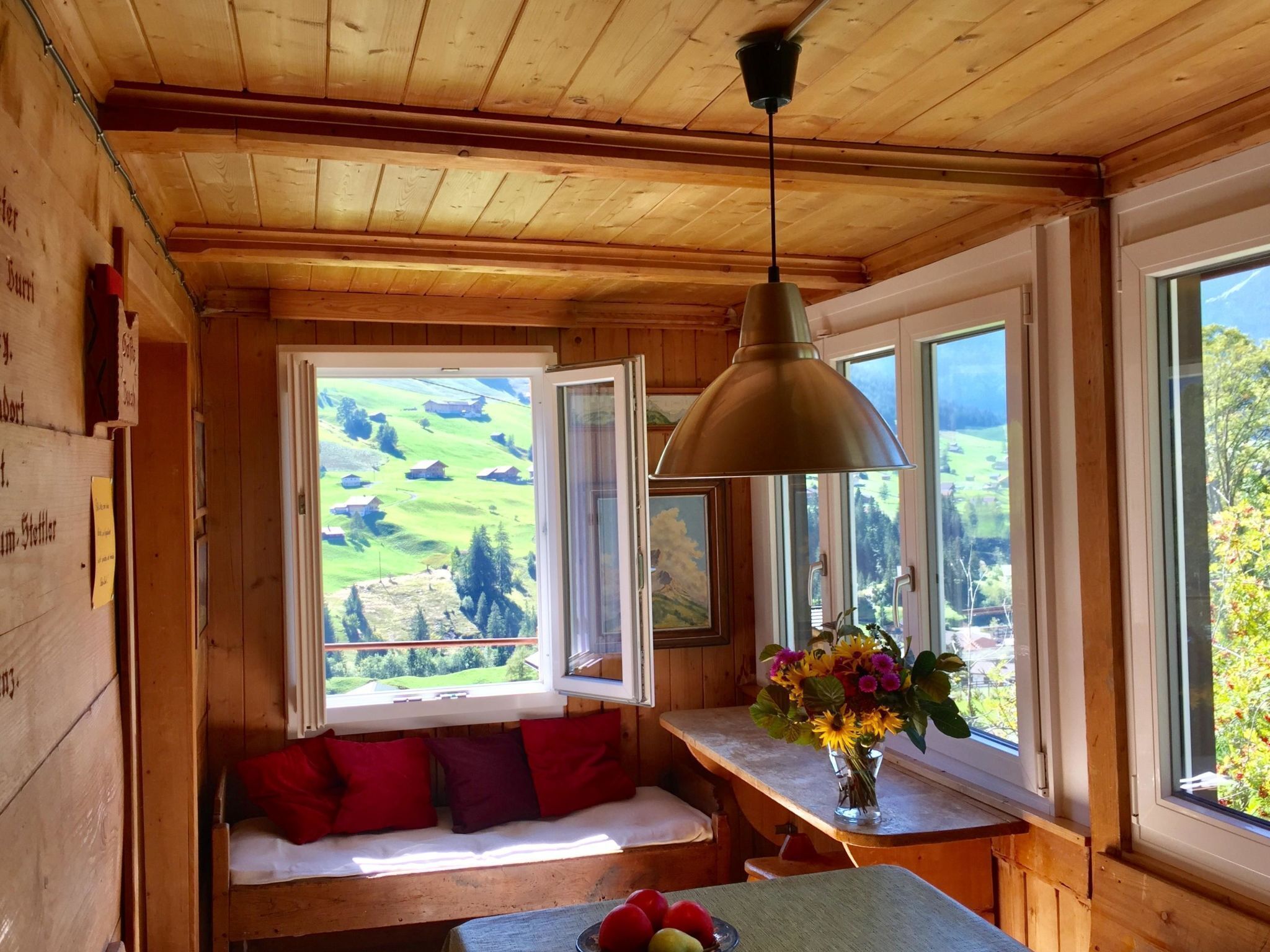 A wooden interior with a bench, a table with fruit, and a view of green fields through windows.