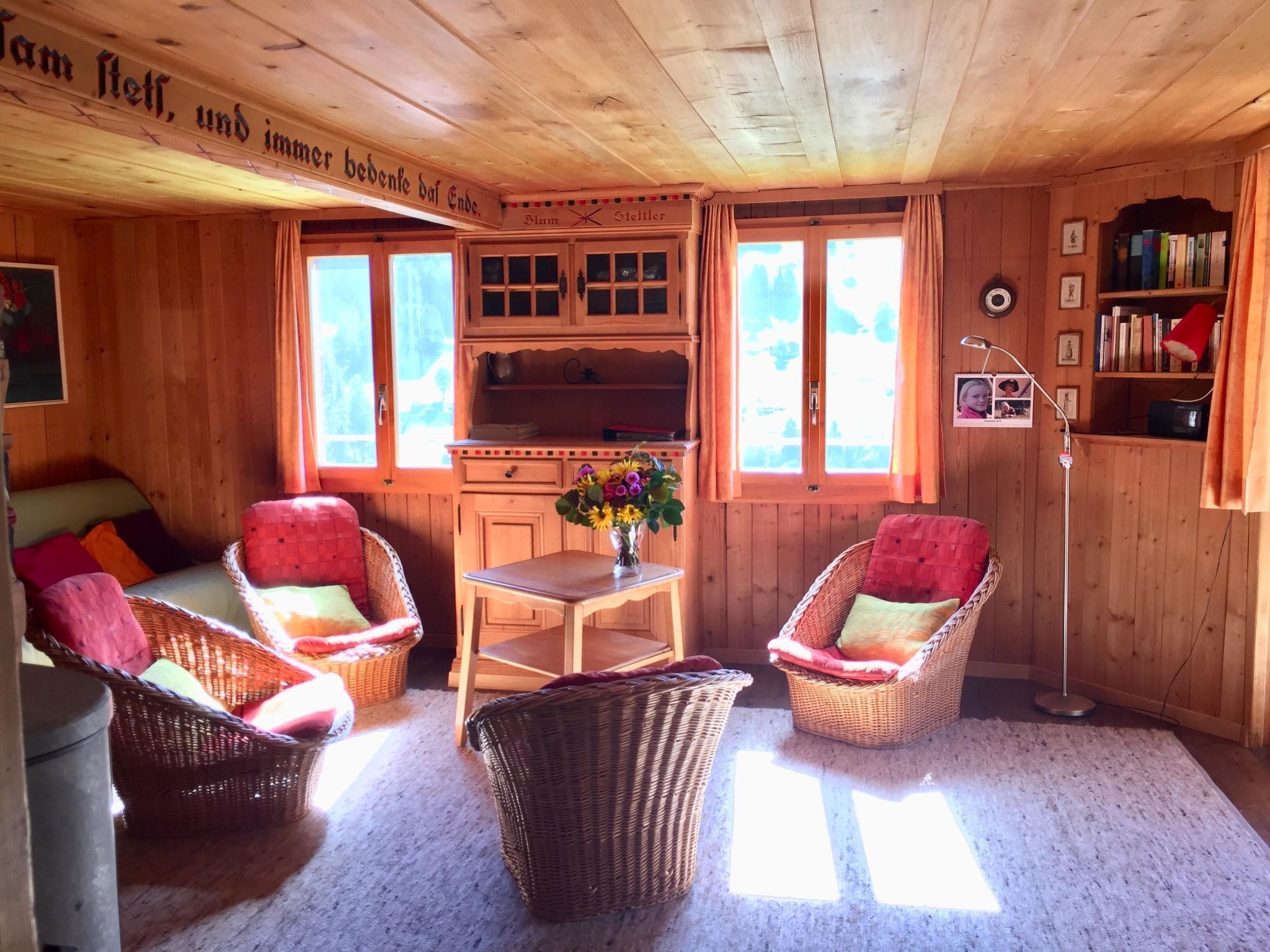 A cozy wooden cabin interior with wicker furniture, a table with a vase of flowers, and a bookshelf.