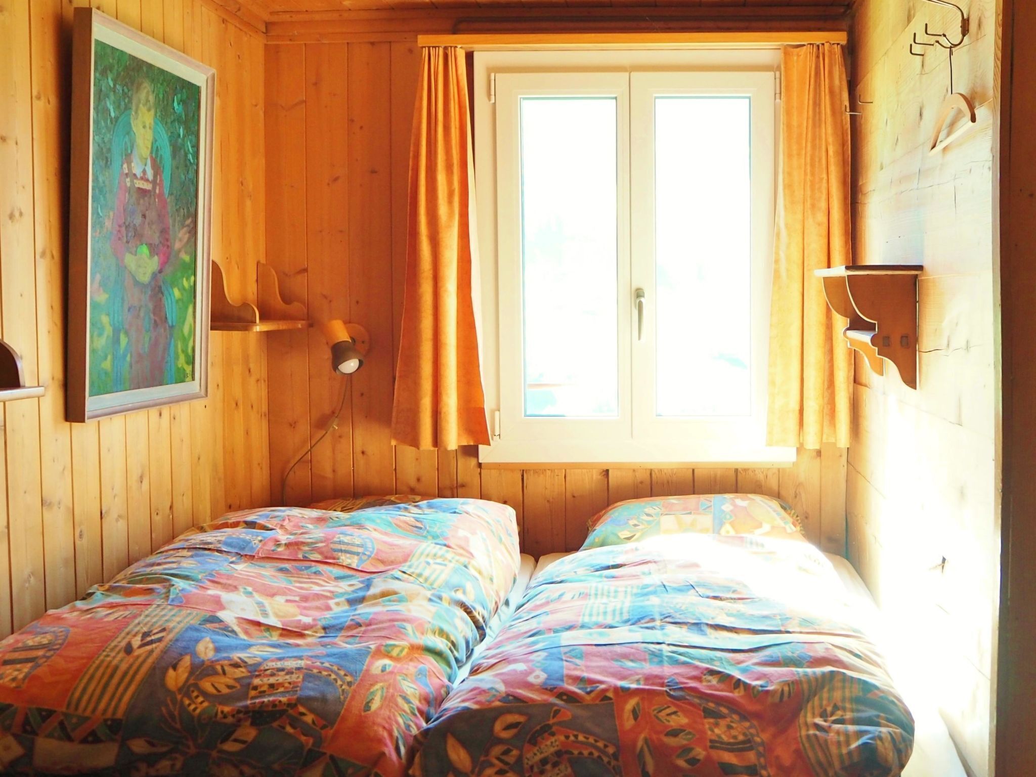 Een kamer met houten wanden, een bed met kleurrijk beddengoed, een raam met gordijnen en een wandlamp.