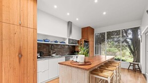 A modern kitchen with wooden cabinets, a central island with bar stools, and a large window offering a view of the outdoors. - Willowbank Homestead (Greater Geelong)