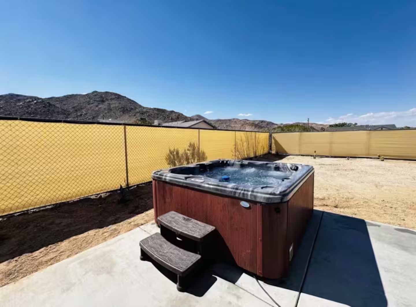 A hot tub in an outdoor setting with a clear sky, surrounded by a yellow fence and a mountain in the background.
