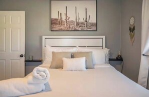 A neatly made bed with white linens and a rolled towel, a wall-mounted picture of cacti, and a dreamcatcher decoration. - 2 bedroom Joshua Tree Retreat with Hot Tub + Cowboy Pool (Twentynine Palms)