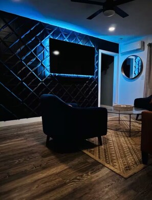 A modern living room with a flat-screen TV mounted on a geometric-patterned wall, a dark armchair, a round mirror, and a dining area with a patterned rug. - 2 bedroom Joshua Tree Retreat with Hot Tub + Cowboy Pool (Twentynine Palms)