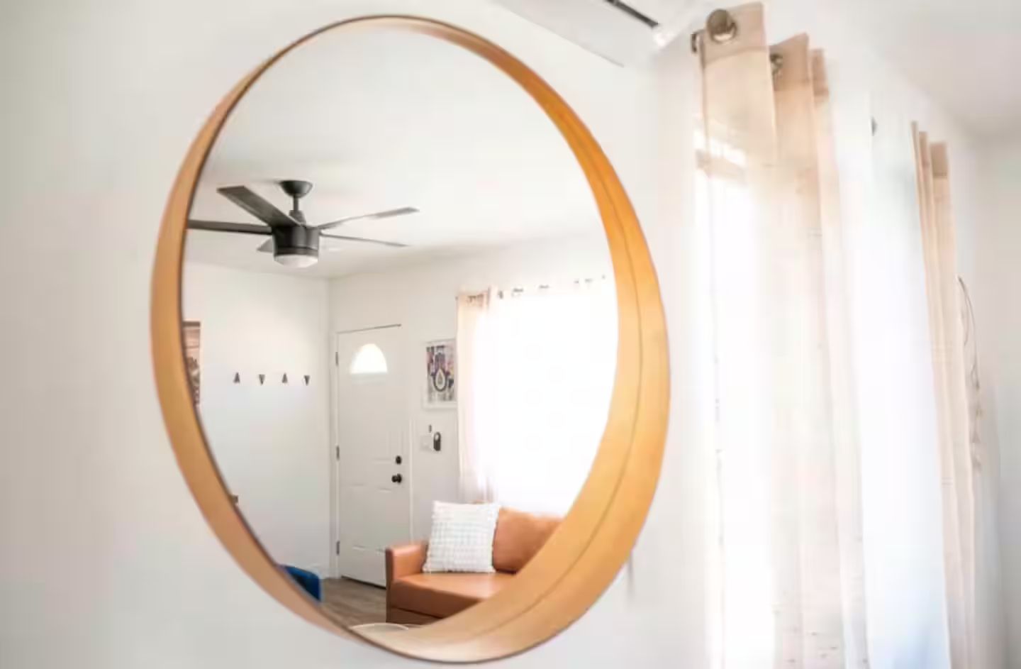 A room with a round mirror on the wall, a ceiling fan, a door, and a window with curtains.