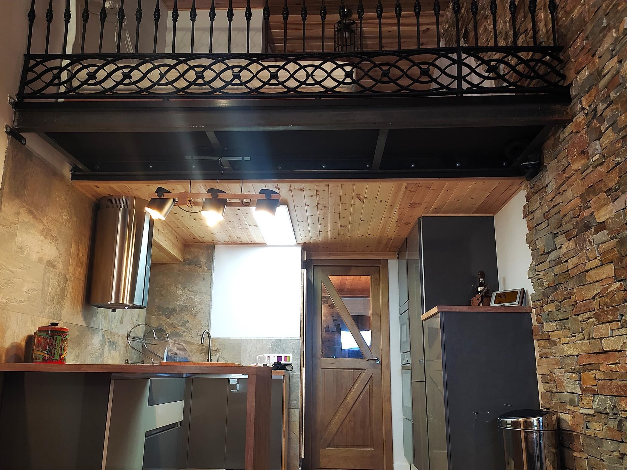Une cuisine avec un plafond en bois, une rambarde en métal noir et un mur en pierre.