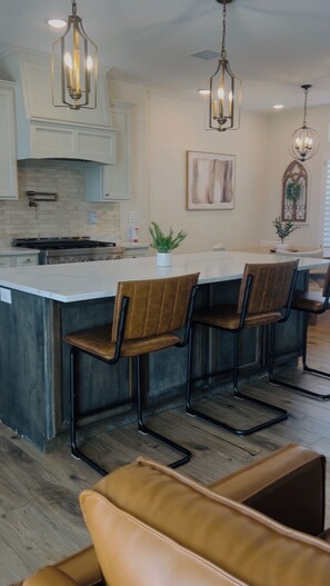 A kitchen with a central island, bar stools, pendant lighting, and a dining area with a sofa.