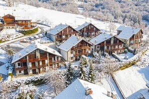 Eine Gruppe von Chaletgebäuden mit mehreren Balkonen, umgeben von schneebedeckten Bäumen und einer Skipiste im Hintergrund.
