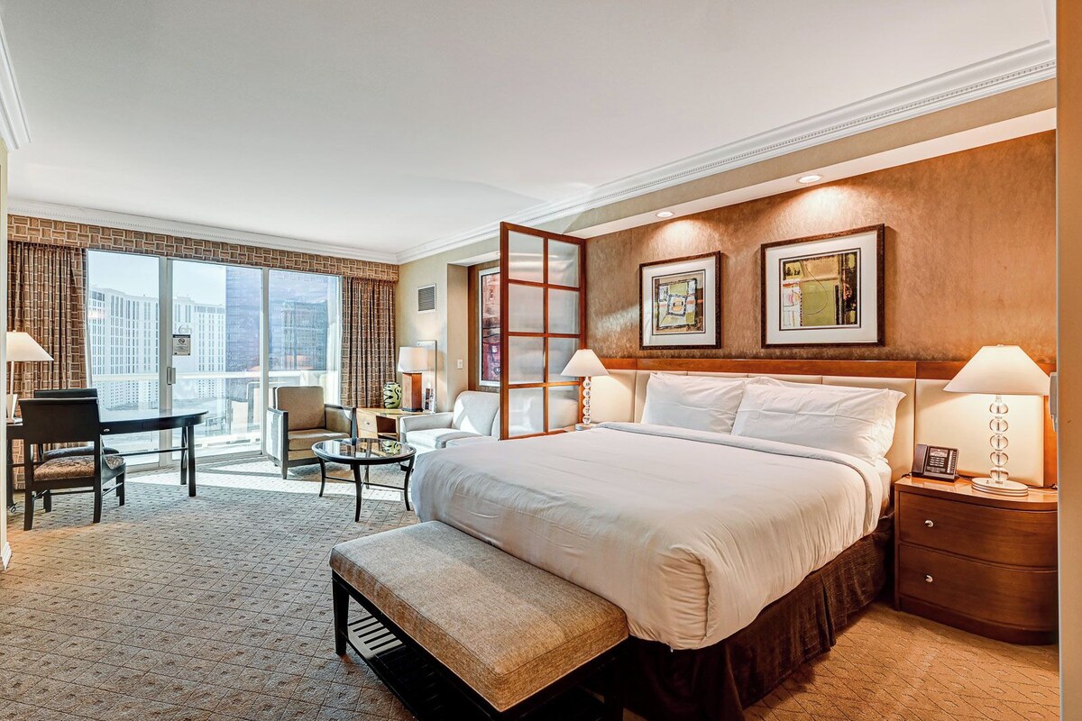 Quarto de hotel com cama grande, escrivaninha, cadeira e varanda com vista para a cidade.