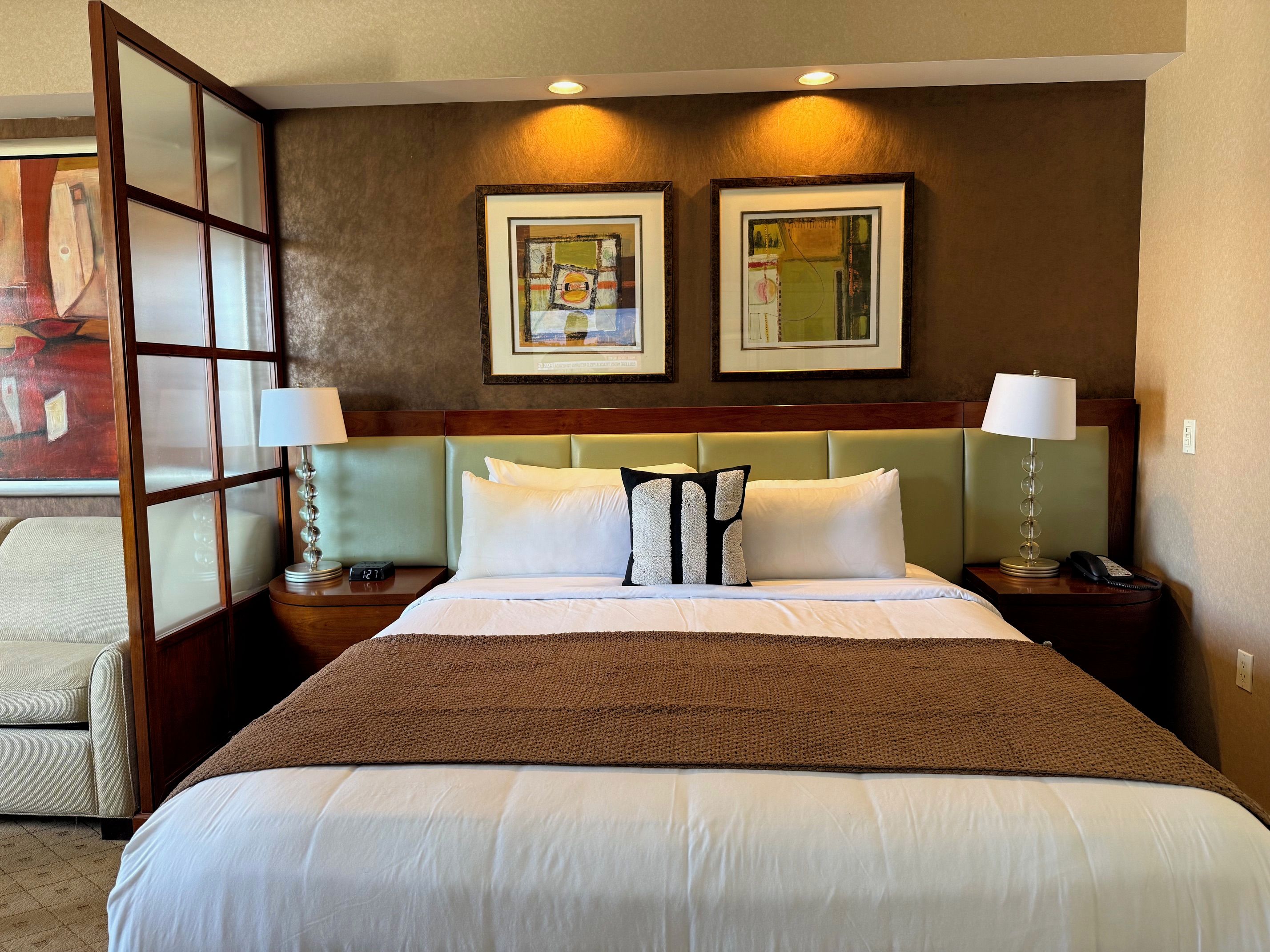 A hotel room with a large bed, two bedside tables with lamps, a headboard, and two framed artworks on the wall.