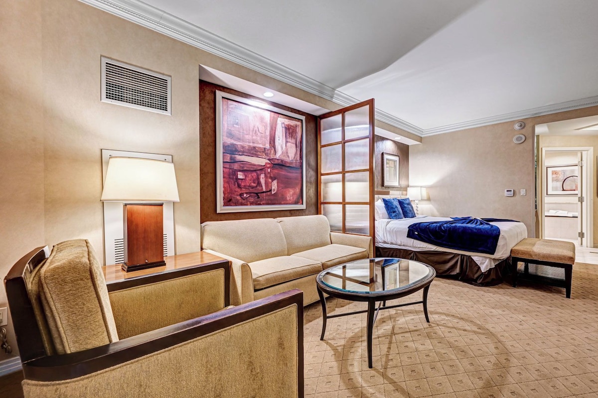 A hotel room with a sofa, a glass coffee table, a bed with a blue blanket, a painting on the wall, and a lamp.