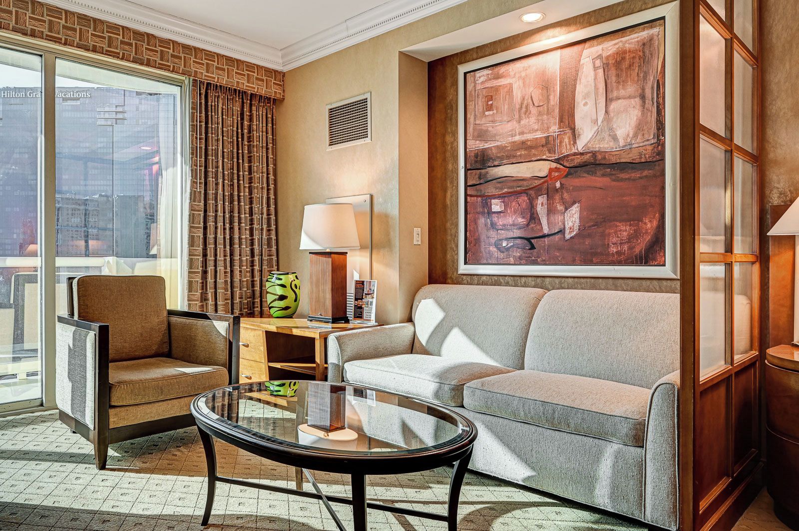 Ein Hotelzimmer mit Sofa, Ohrensessel und einem Glastisch. An der Wand hängt ein großes abstraktes Gemälde und ein Fenster mit Vorhängen.