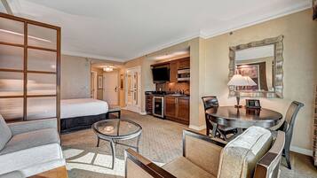 Habitación de hotel con cama, sofá, mesa de centro de vidrio, mesa de comedor con sillas, zona de cocina y un espejo grande.