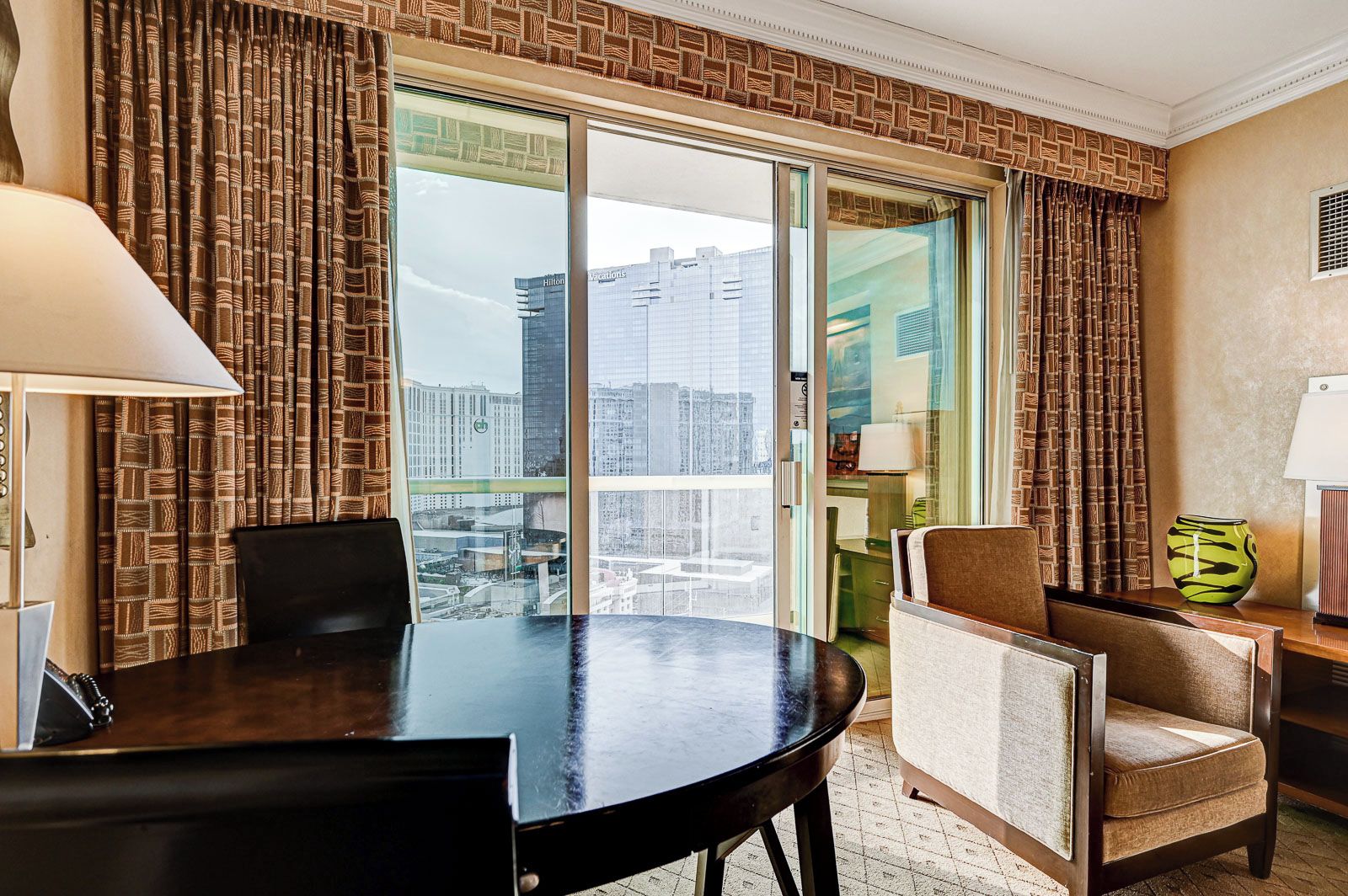 Une chambre d’hôtel dotée d’une grande porte-fenêtre coulissante donnant sur un balcon avec vue sur la ville. On y trouve une table ronde avec deux chaises, une petite table d’appoint avec une lampe et un vase décoratif.