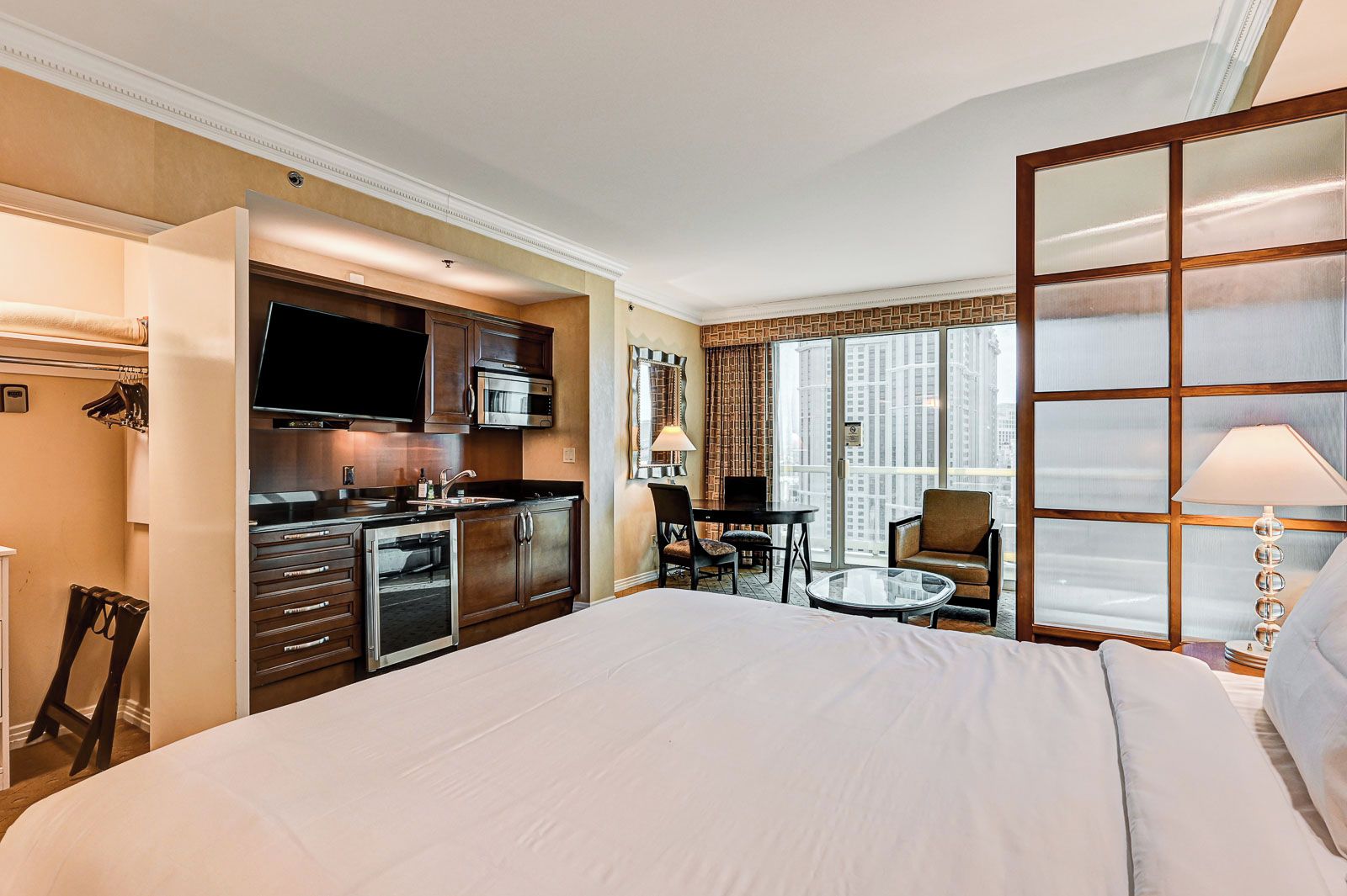 A hotel room with a large bed, kitchenette, dining area, and a view of the city.