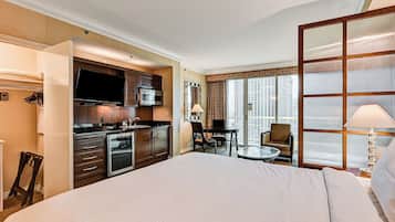 A hotel room with a large bed, kitchenette, dining area, and a view of the city.