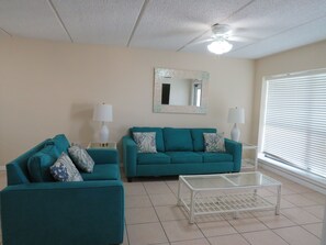A living room with teal sofas, a glass coffee table, two lamps, a ceiling fan, and a large window with blinds. - SPI Waterfront Getaway - Wake up to Ocean Views (South Padre Island)