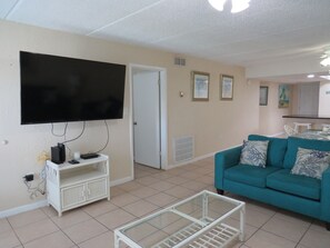 Ein Wohnzimmer mit Flachbildfernseher, einem türkisblauen Sofa, einem Glastisch und einer weißen Unterhaltungsecke.