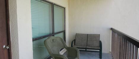 A balcony with a wicker chair, a cushioned bench, and a closed window with blinds.