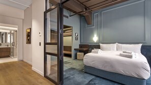 Ein modernes Hotelzimmer mit einem großen Bett, einer Glastür zu einem Badezimmer und Blick auf einen Flur.