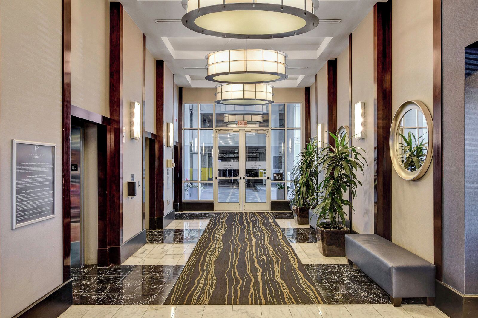 A modern hotel lobby with a marble floor, a bench, potted plants, and a large glass door leading to the reception area.