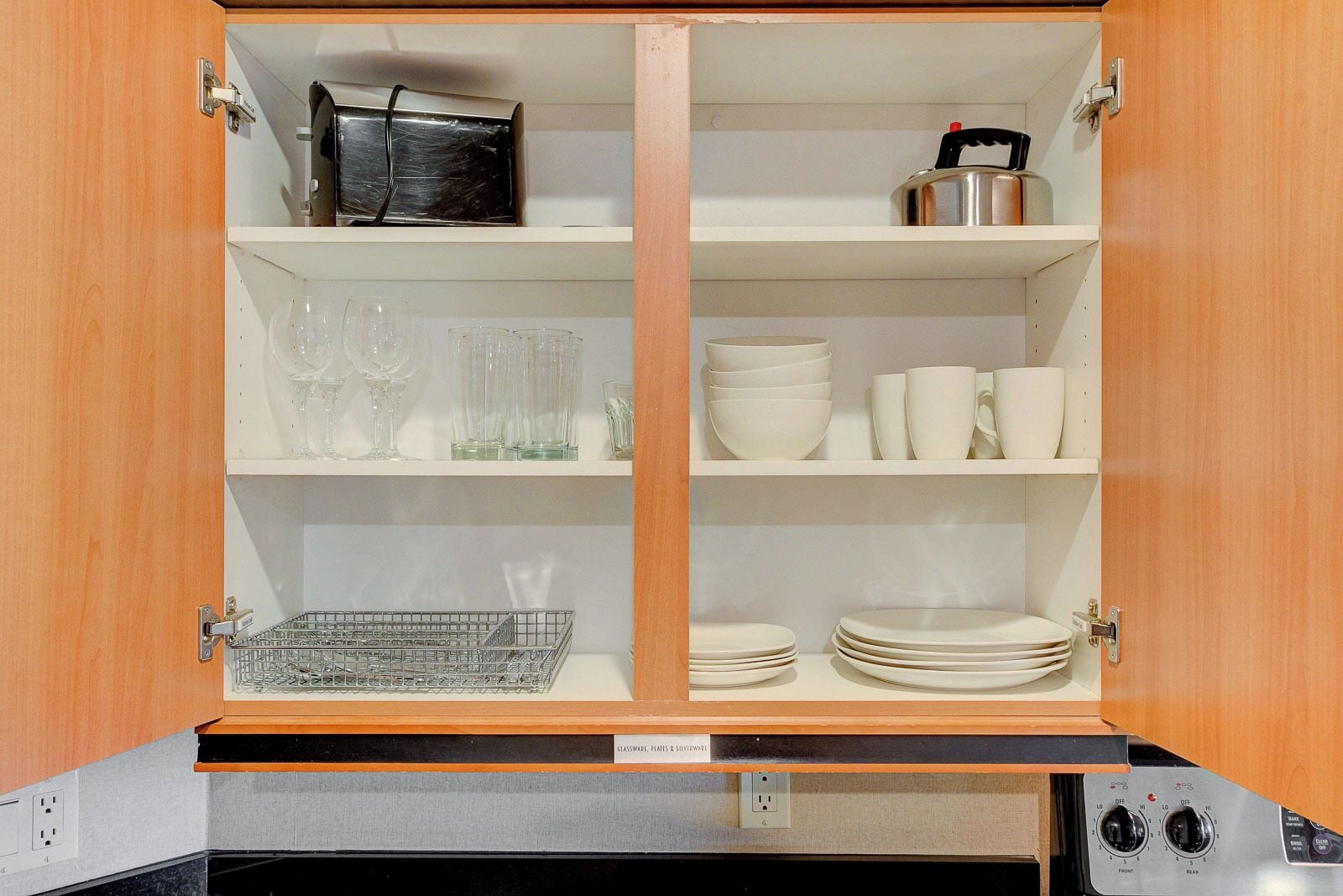 A kitchen cabinet with shelves containing glassware, plates, and a kettle.