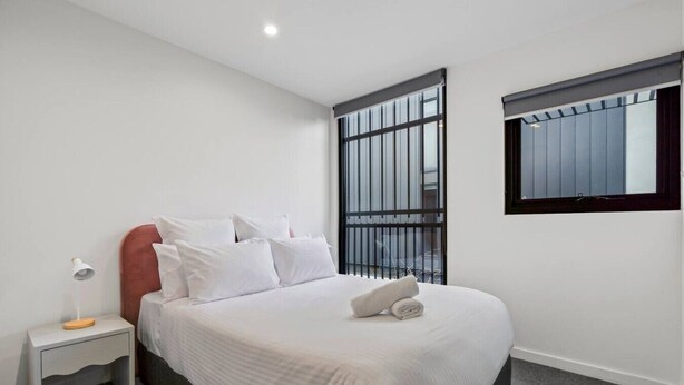 A bedroom with a bed, a nightstand, a lamp, and a window. - Leafy Caulfield Townhouse - (pool, car park, BBQ) (Caulfield North)