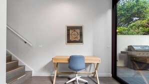 A room with a wooden desk, a chair, a staircase, and a view of an outdoor grill through a glass door. - Leafy Caulfield Townhouse - (pool, car park, BBQ) (Caulfield North)
