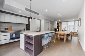A modern kitchen with a central island, bar stools, and a dining area with a wooden table and chairs. - Leafy Caulfield Townhouse - (pool, car park, BBQ) (Caulfield North)