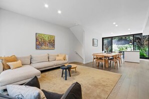 A modern living space with a sofa, coffee table, dining table, and chairs. There is a painting on the wall and a window providing a view of the outdoors. - Leafy Caulfield Townhouse - (pool, car park, BBQ) (Caulfield North)