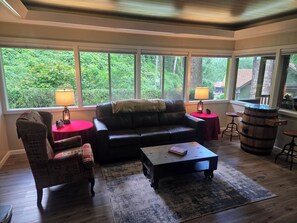 A living room with a leather sofa, two armchairs, a coffee table, and a rug. There are two lamps on side tables, and a wooden barrel. Large windows provide a view of greenery outside. - Mountain Memories Gatlinburg Home Sleeps 9 NO Winding Roads (Gatlinburg)