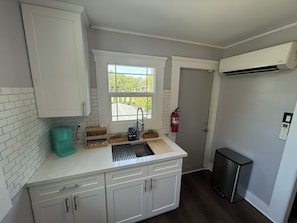 A kitchen with white cabinets, a window, a sink, a fire extinguisher, and a refrigerator. - Welcome to the Aviator - 1920's Craftsman Airplane Bungalow (Modesto)