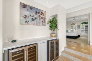 A modern interior with a white wall, a framed artwork of palm trees, a potted plant, and a minibar with a Vintec logo. - Tranquillity - Poolside Sanctuary & Rainforest Views (Smithfield)
