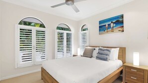 A bedroom with a bed, a nightstand, a ceiling fan, and a painting of a beach scene. - Tranquillity - Poolside Sanctuary & Rainforest Views (Smithfield)