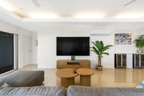 A modern living room with a flat-screen TV, a sofa, a coffee table, and a potted plant. - Tranquillity - Poolside Sanctuary & Rainforest Views (Smithfield)