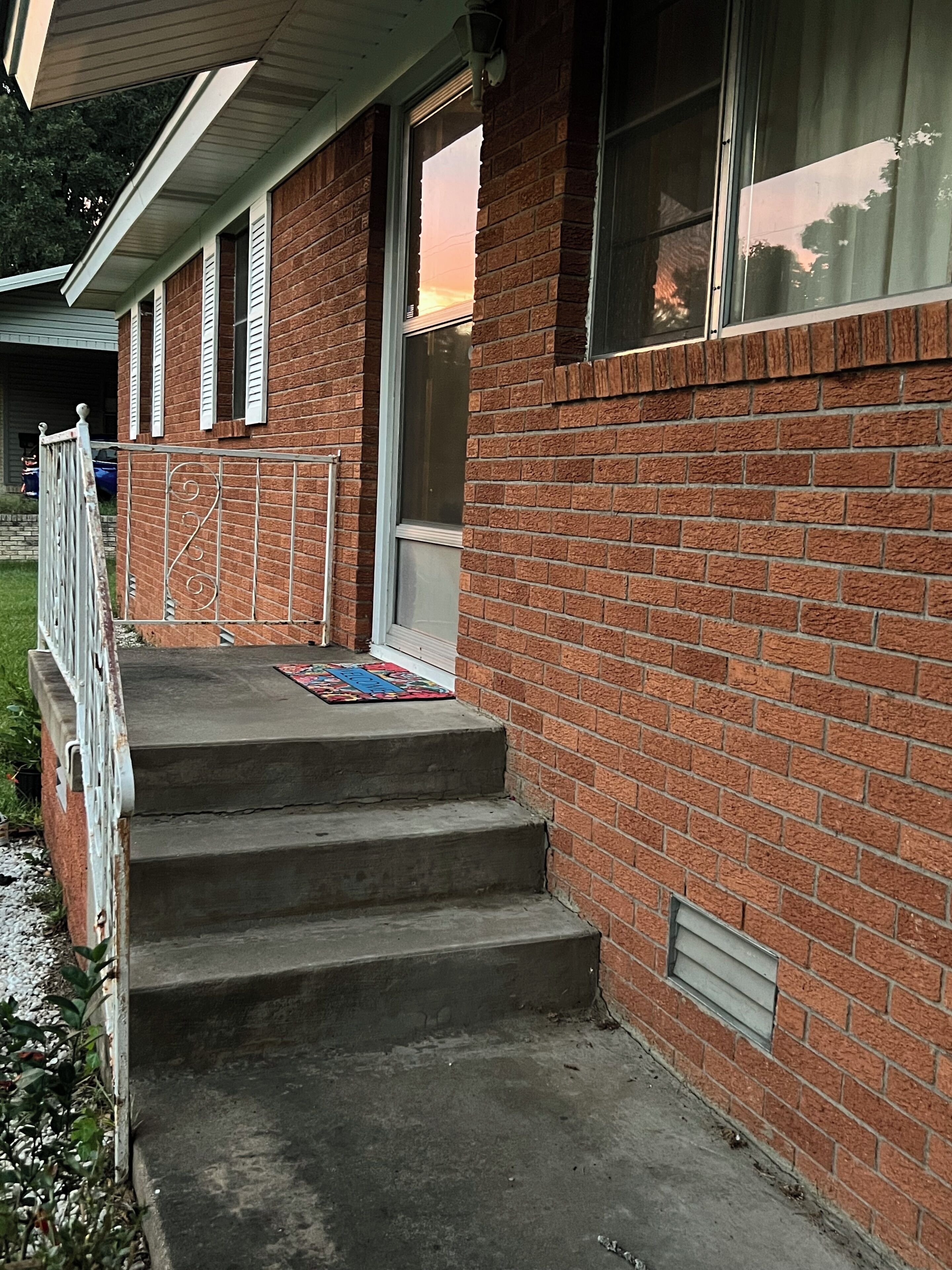 A brick building with a set of concrete steps leading to a door, a metal railing, and a window with blinds.