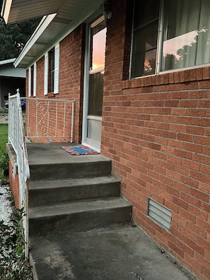 A brick building with a set of concrete steps leading to a door, a metal railing, and a window with blinds.