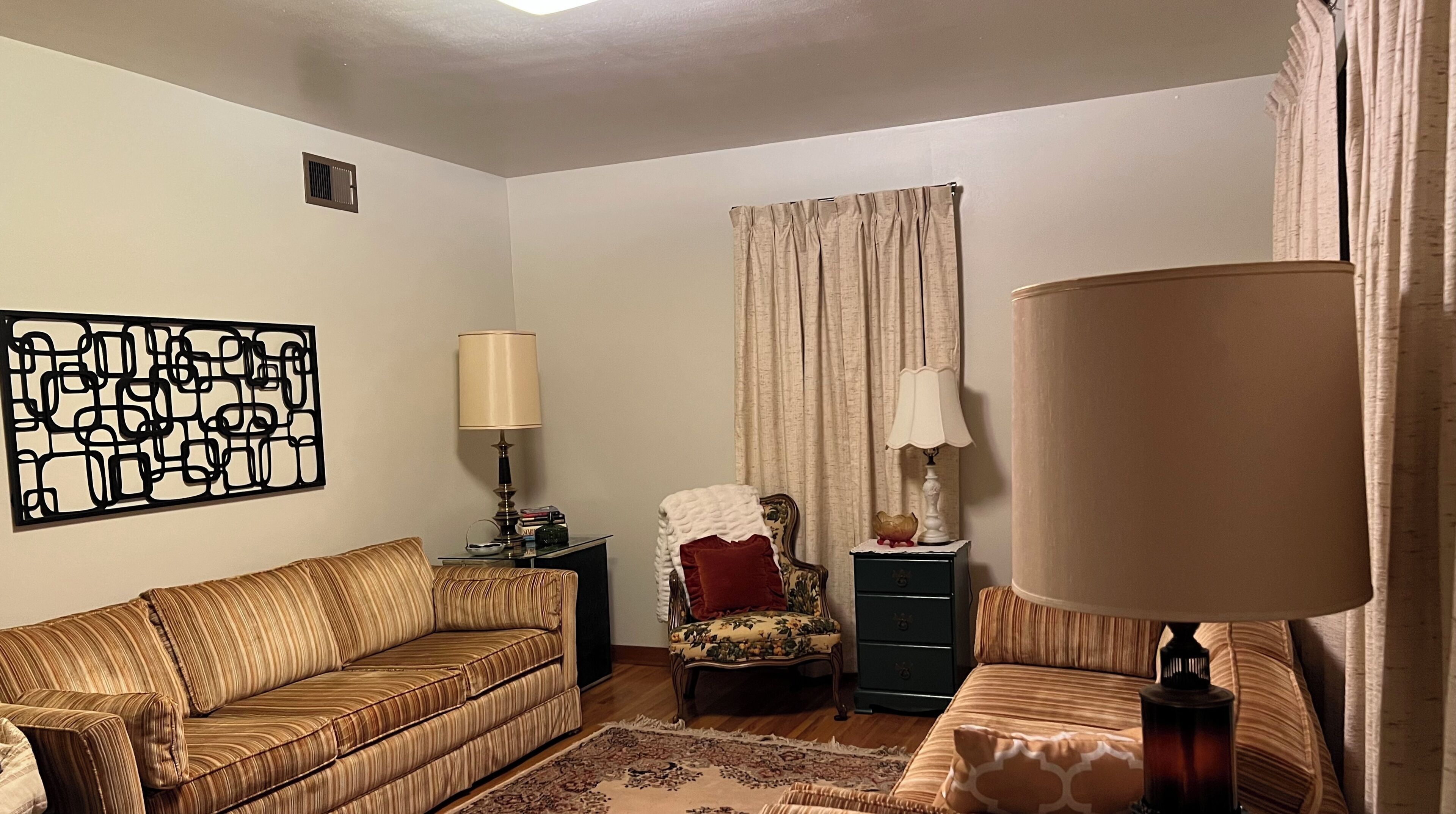 A living room with a sofa, a chair, a lamp, a decorative wall piece, and a window with curtains.