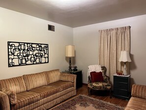A living room with a striped sofa, a patterned armchair, a small table with a lamp, a decorative rug, and a wall art piece.