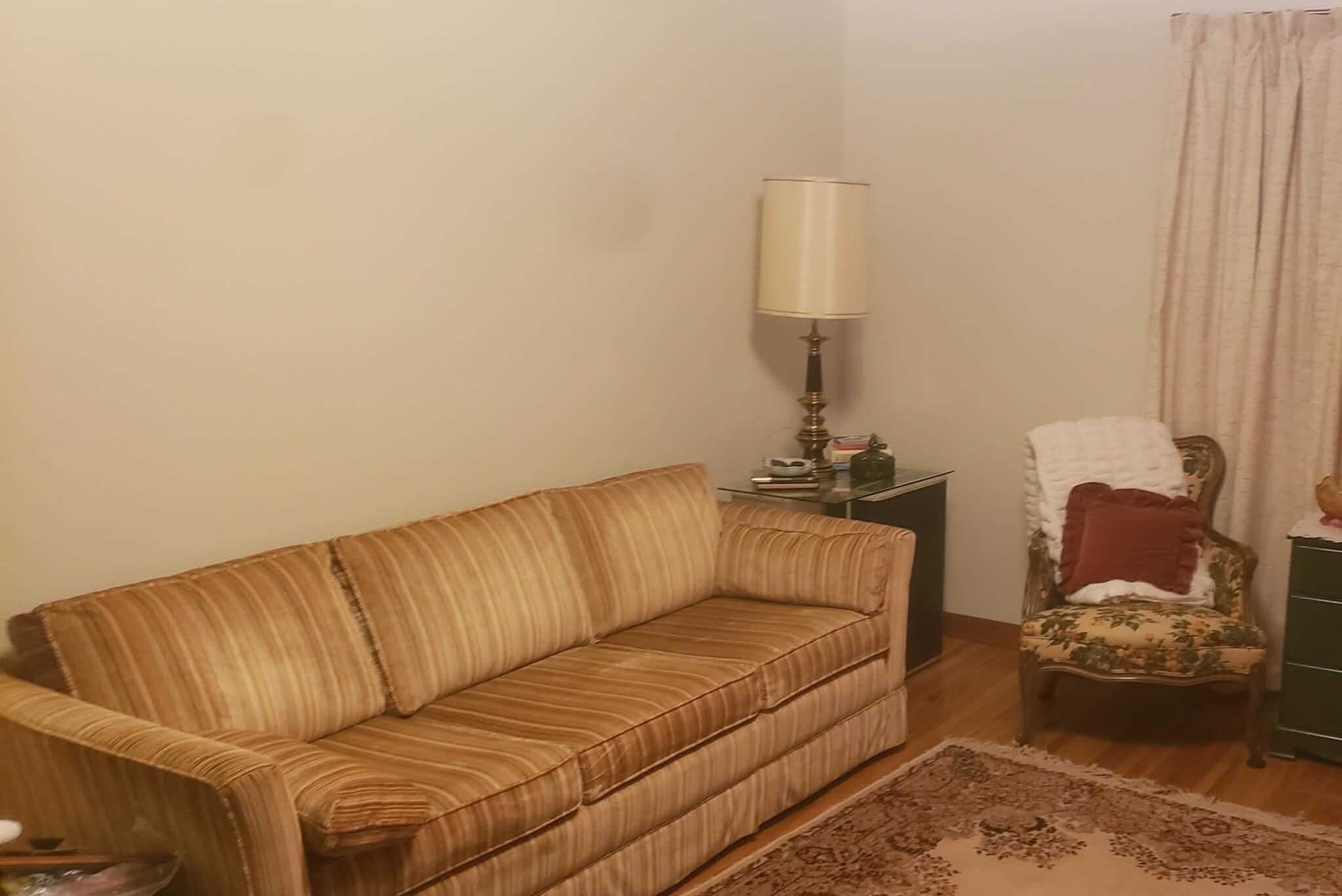 Una sala de estar con un sofá, una mesita de noche con una lámpara, un sillón floral y una alfombra estampada.