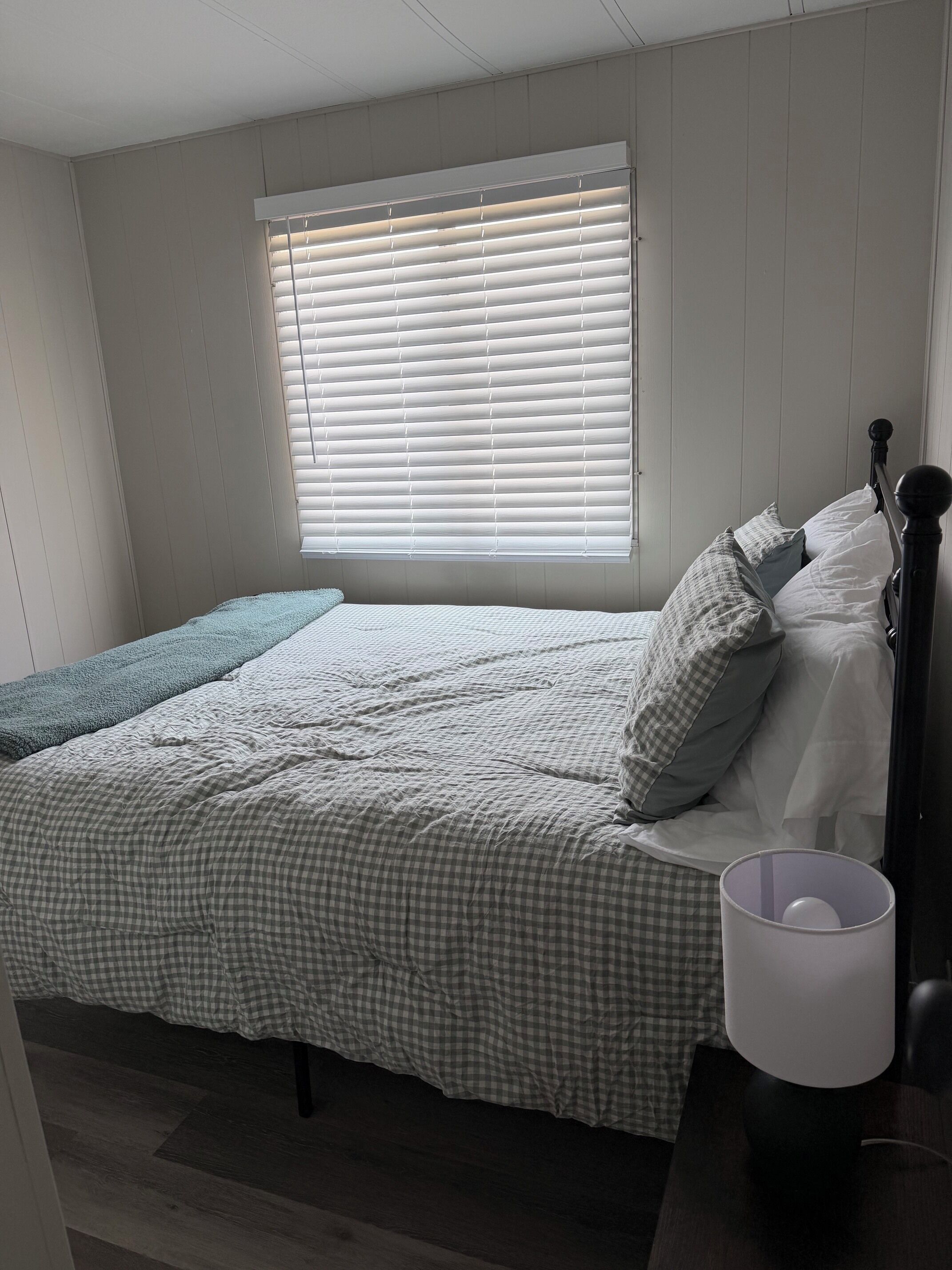 Un dormitorio con cama, almohadas, una lámpara de noche y una ventana con persianas.