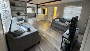 A modern living room with a grey sofa, a TV on a stand, and a kitchen in the background.