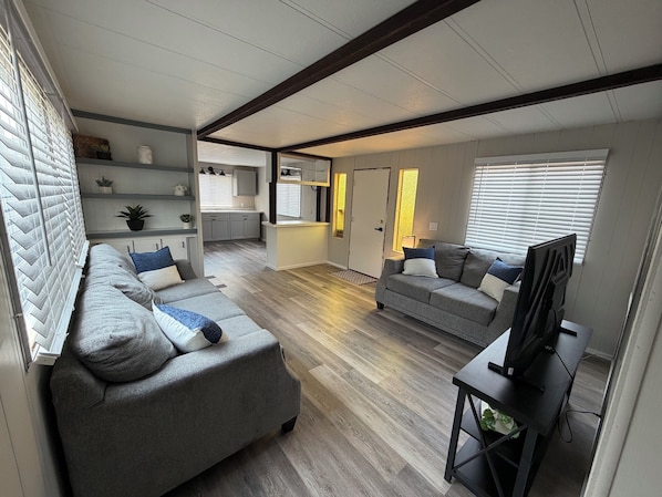 A modern living room with a grey sofa, a TV on a stand, and a kitchen in the background.