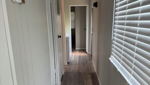 A hallway with wooden flooring, white walls, and a window with blinds.