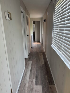 A hallway with wooden flooring, white walls, and a window with blinds.