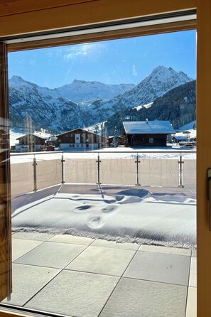 Blick durch ein Fenster auf ein Gebäude mit verschneitem Gelände, einem Holzzaun und einer Berglandschaft im Hintergrund.